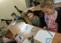 Uczestnicy siedzący w ławkach, przed nimi znajdują się kartki i zestawy geometryczne